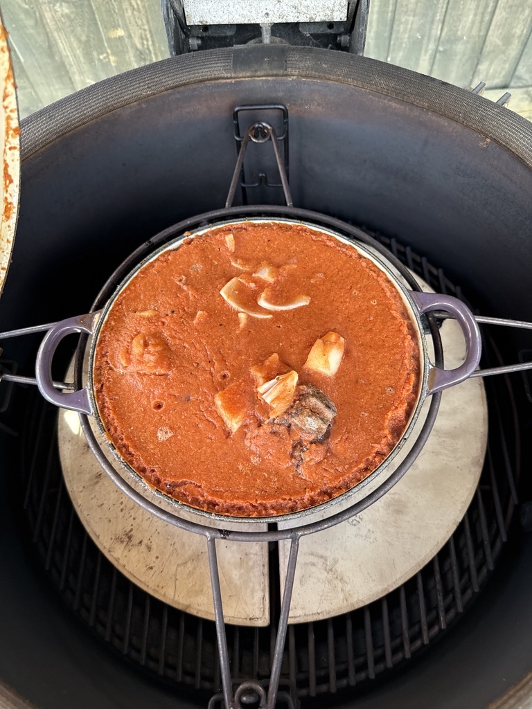 Cast iron pot of birria sauce and beef set inside a Kamado BBQ